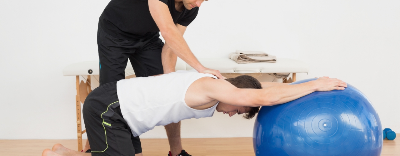 Male patient performing therapeutic exercises with the aide of a body ball and under the guidance of a physical therapsit.